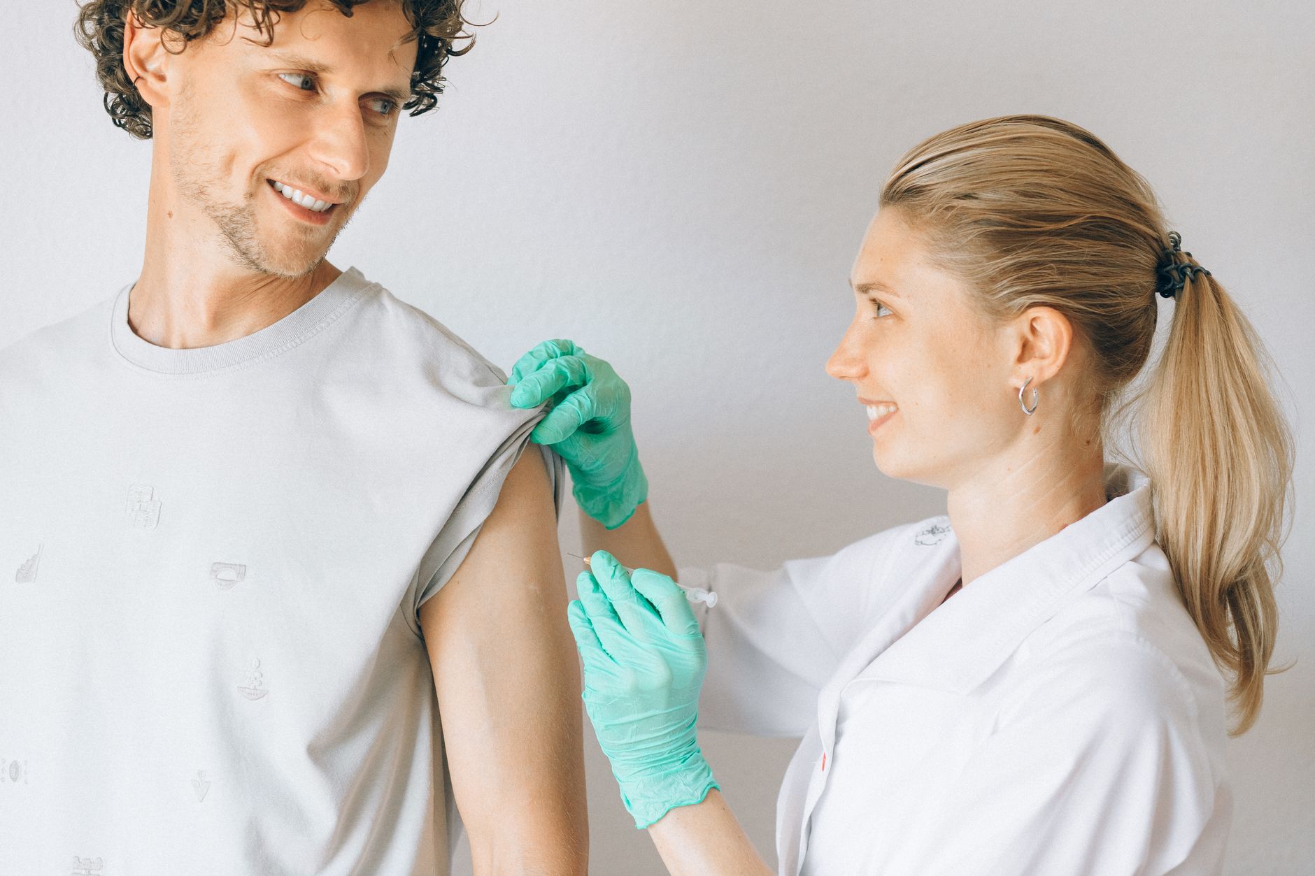 A clinician in green gloves gives an injection in the upper arm of a smiling man who holds up his sleeve
