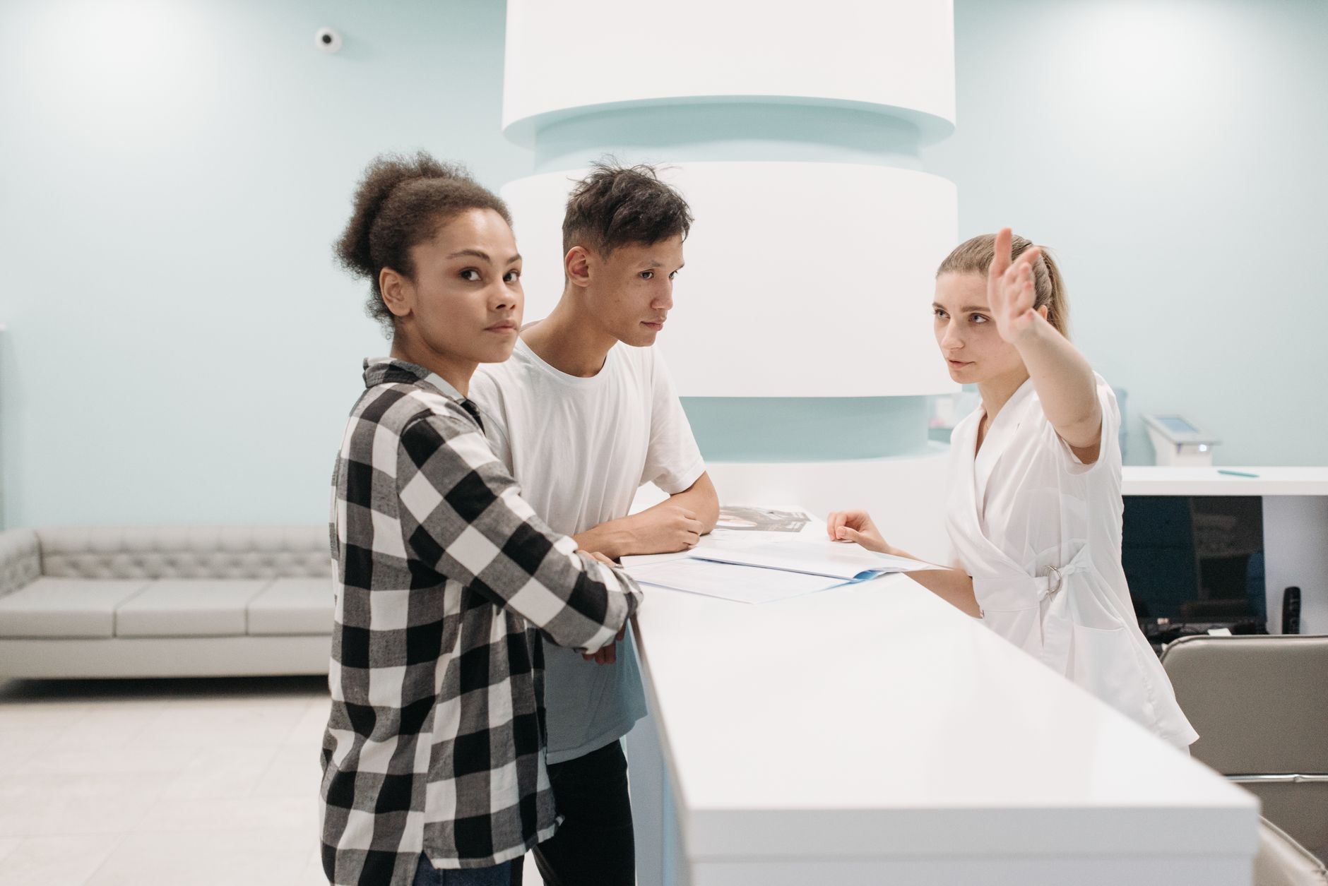 A clinic staff member at a white front desk points the way for two patients checking in with paperwork