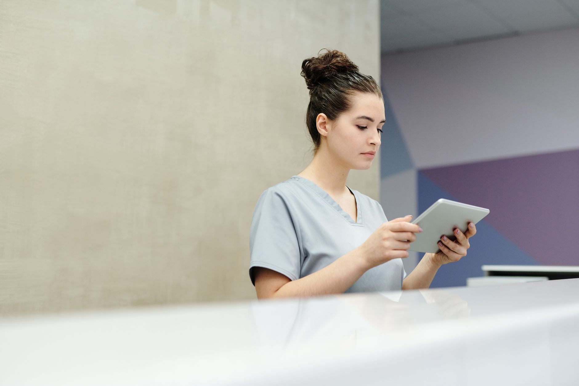 A medical assistant in gray scrubs reads a tablet while standing behind a clinic reception desk