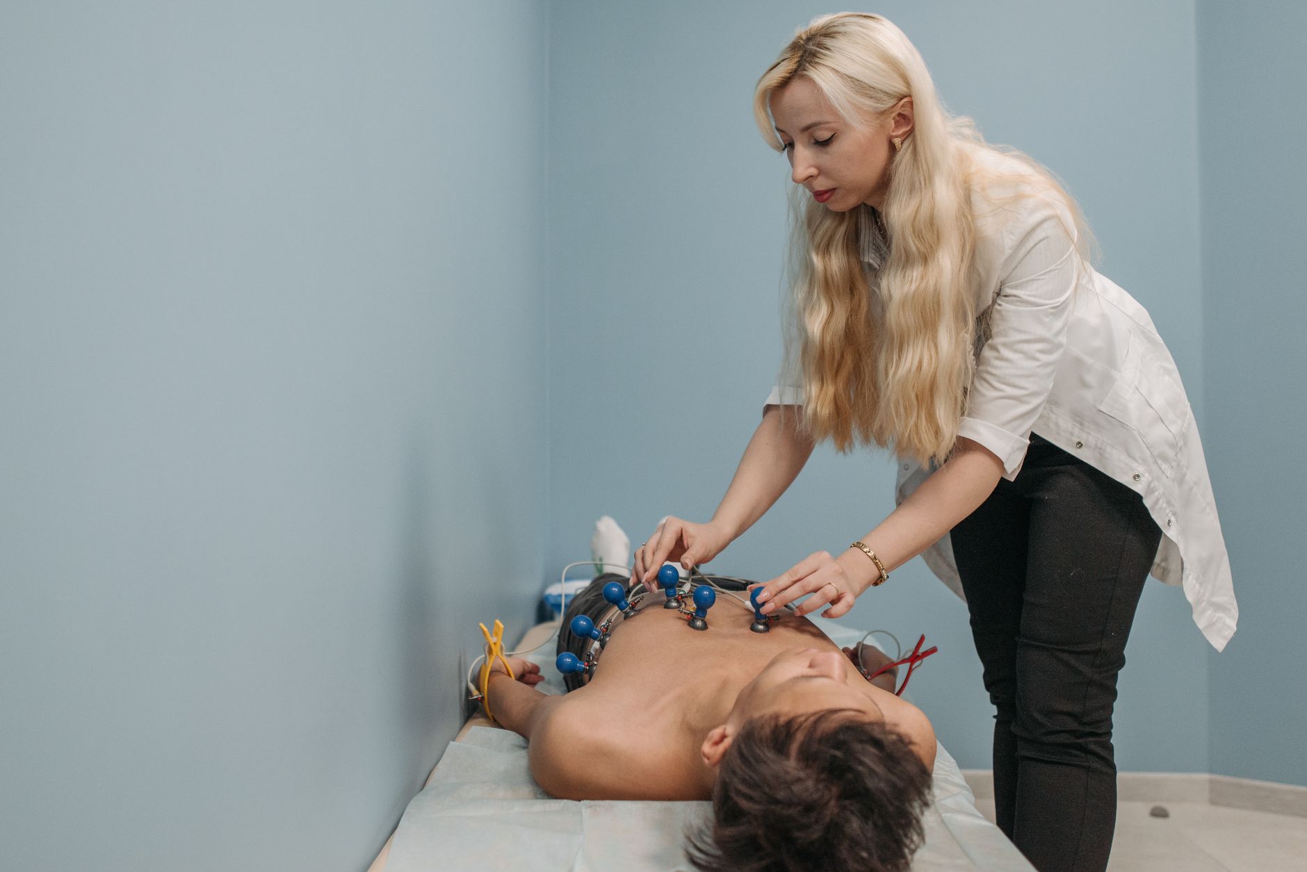 A clinician in a white coat places suction-cup EKG electrodes across a patient's chest on an exam table