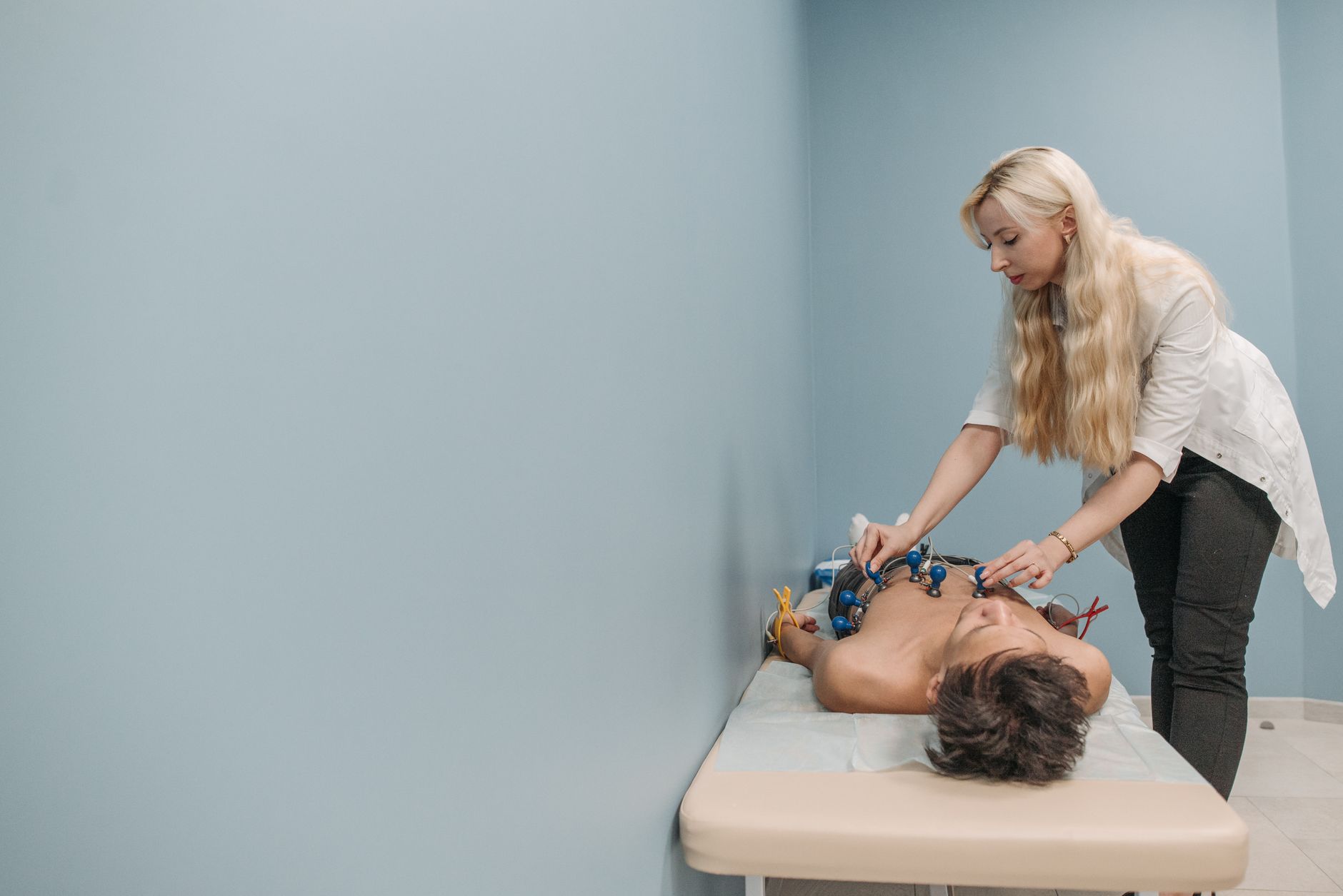 A clinician in a white coat attaches EKG suction electrodes to the chest of a patient lying on an exam table