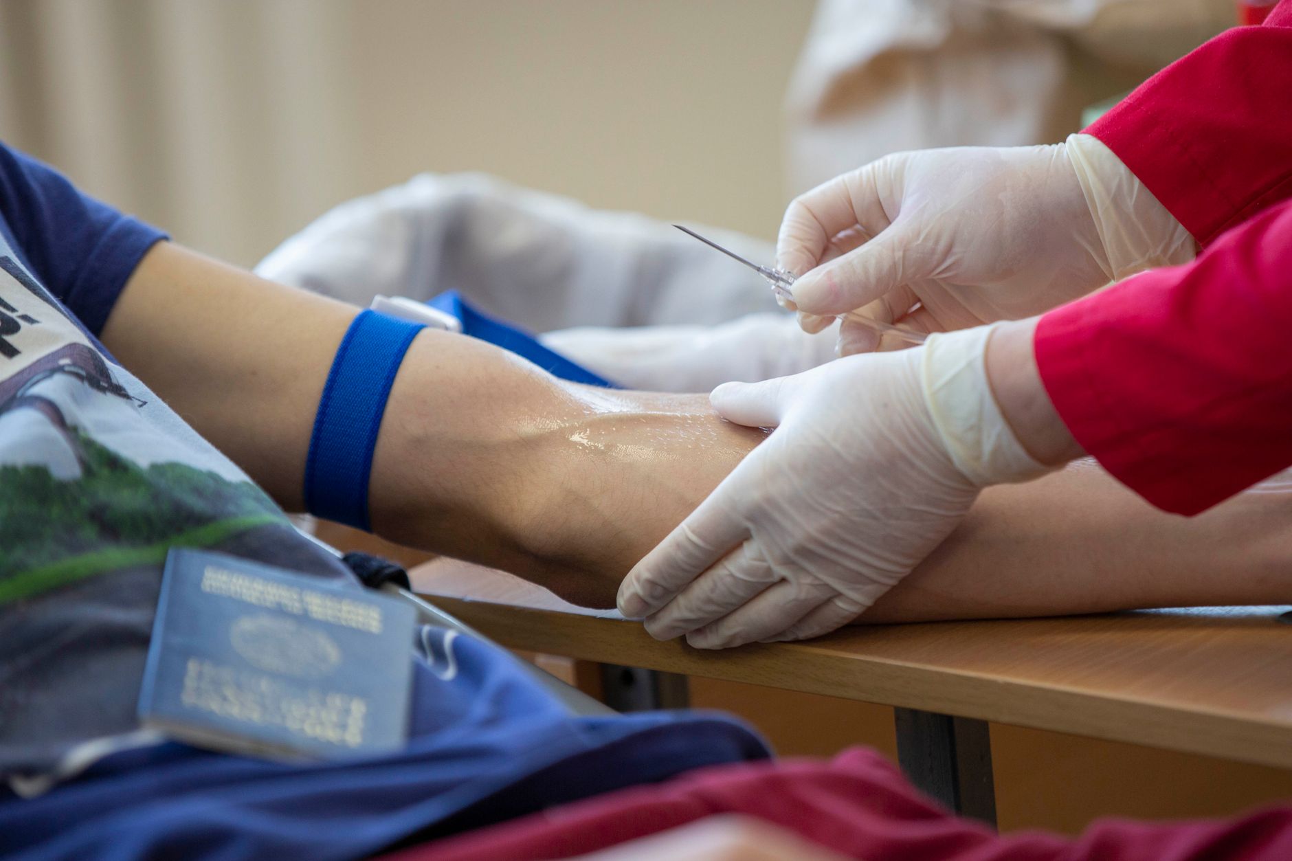 Gloved hands steady a patient's arm below a blue tourniquet and hold a needle ready for a blood draw