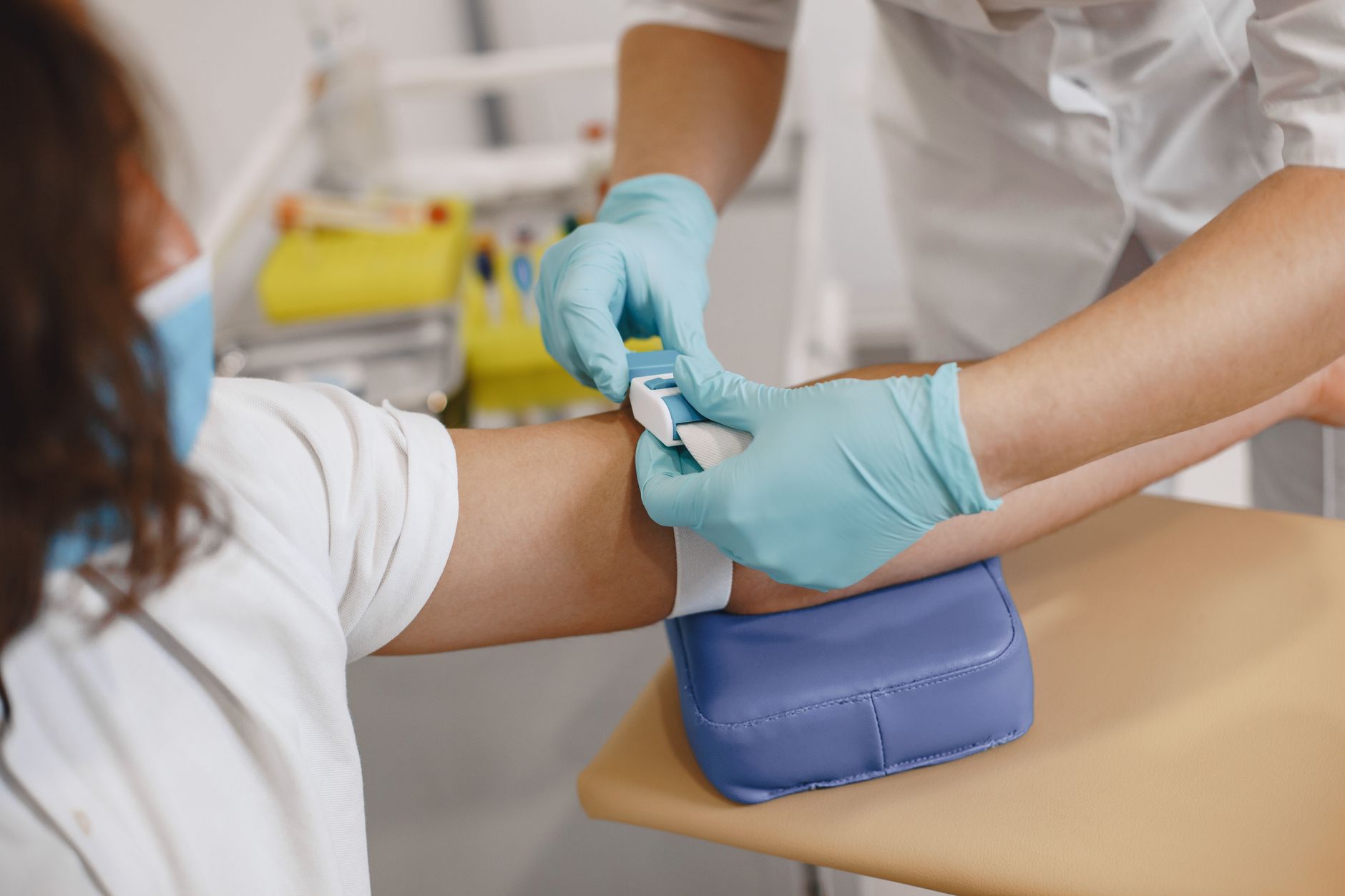 A gloved clinician fastens a tourniquet on a seated patient's upper arm, resting on a padded arm rest, before a blood draw