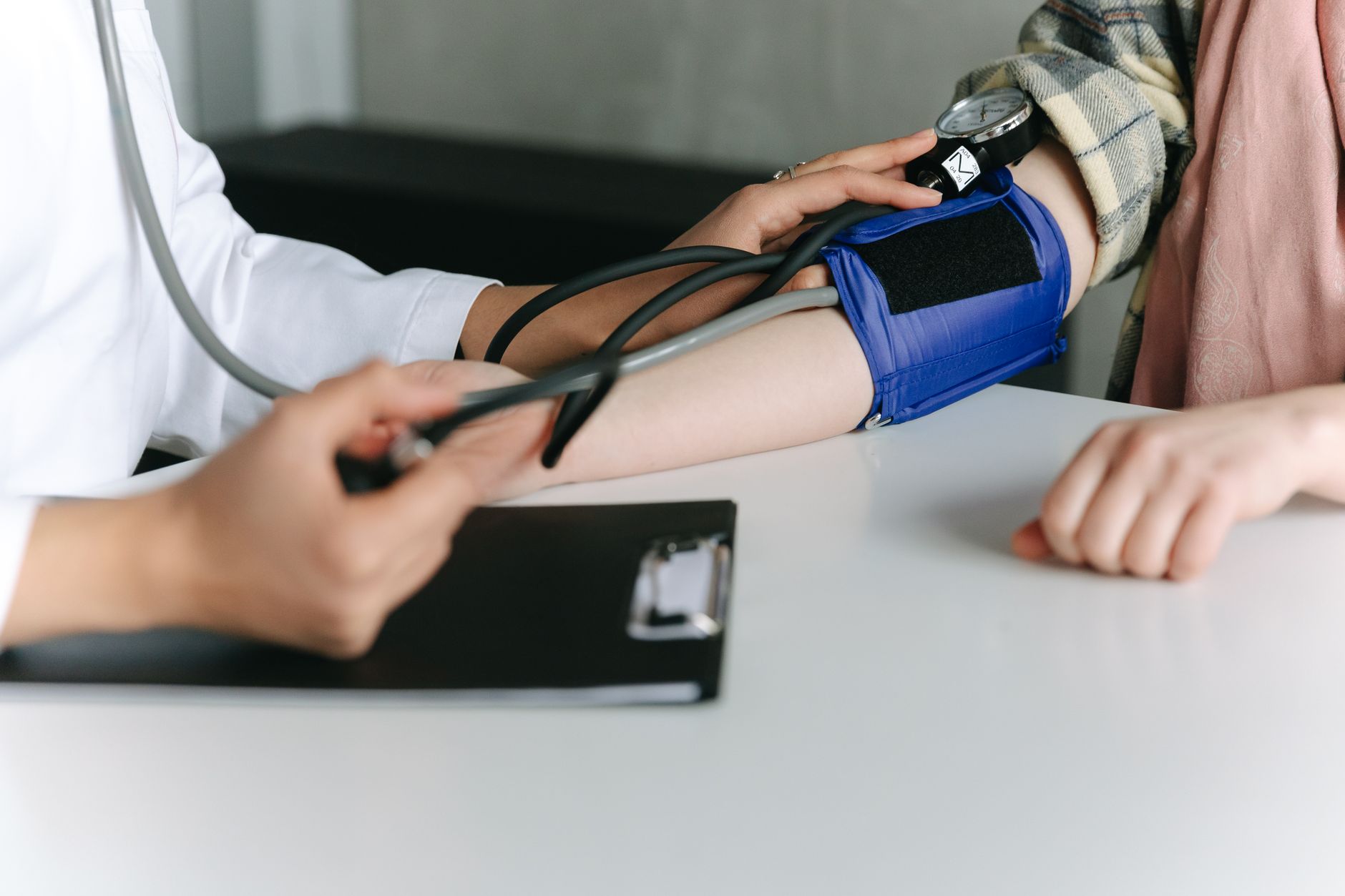 A clinician in a white coat takes a patient's blood pressure with a manual cuff and stethoscope at a desk