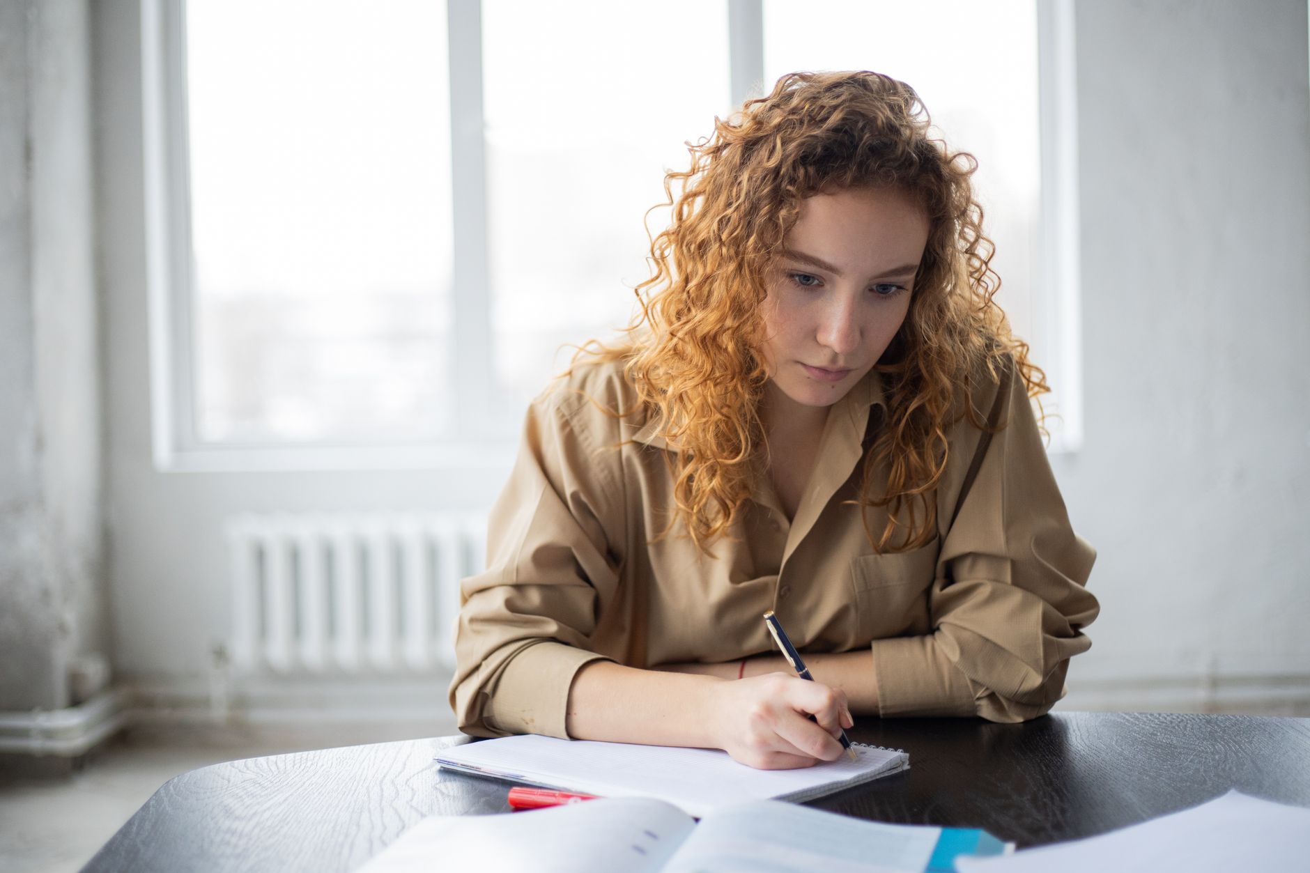 A student with curly hair writes notes in a notebook at a desk beside an open textbook