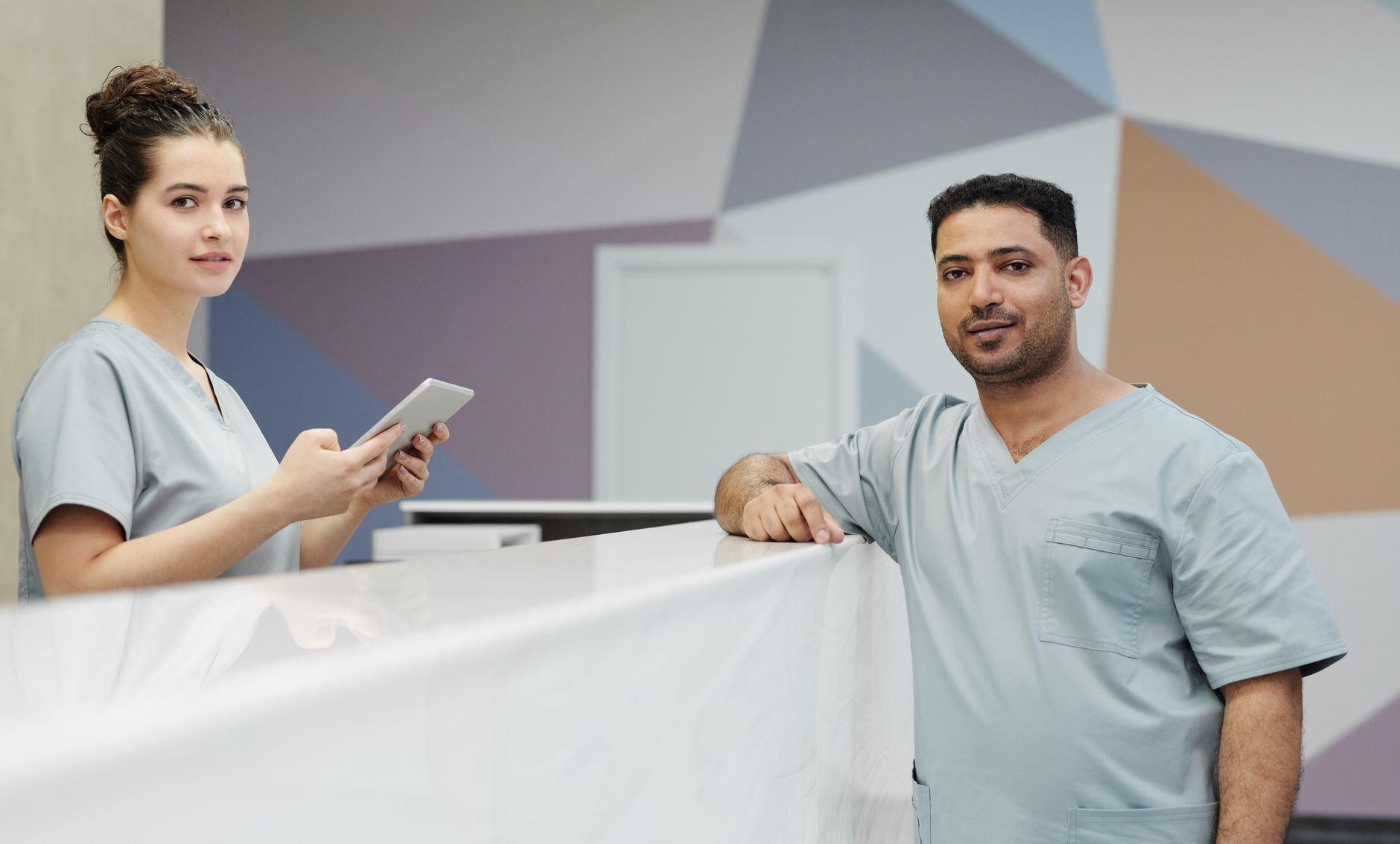 Two clinic staff members in light blue scrubs stand at a medical office front desk, one holding a tablet