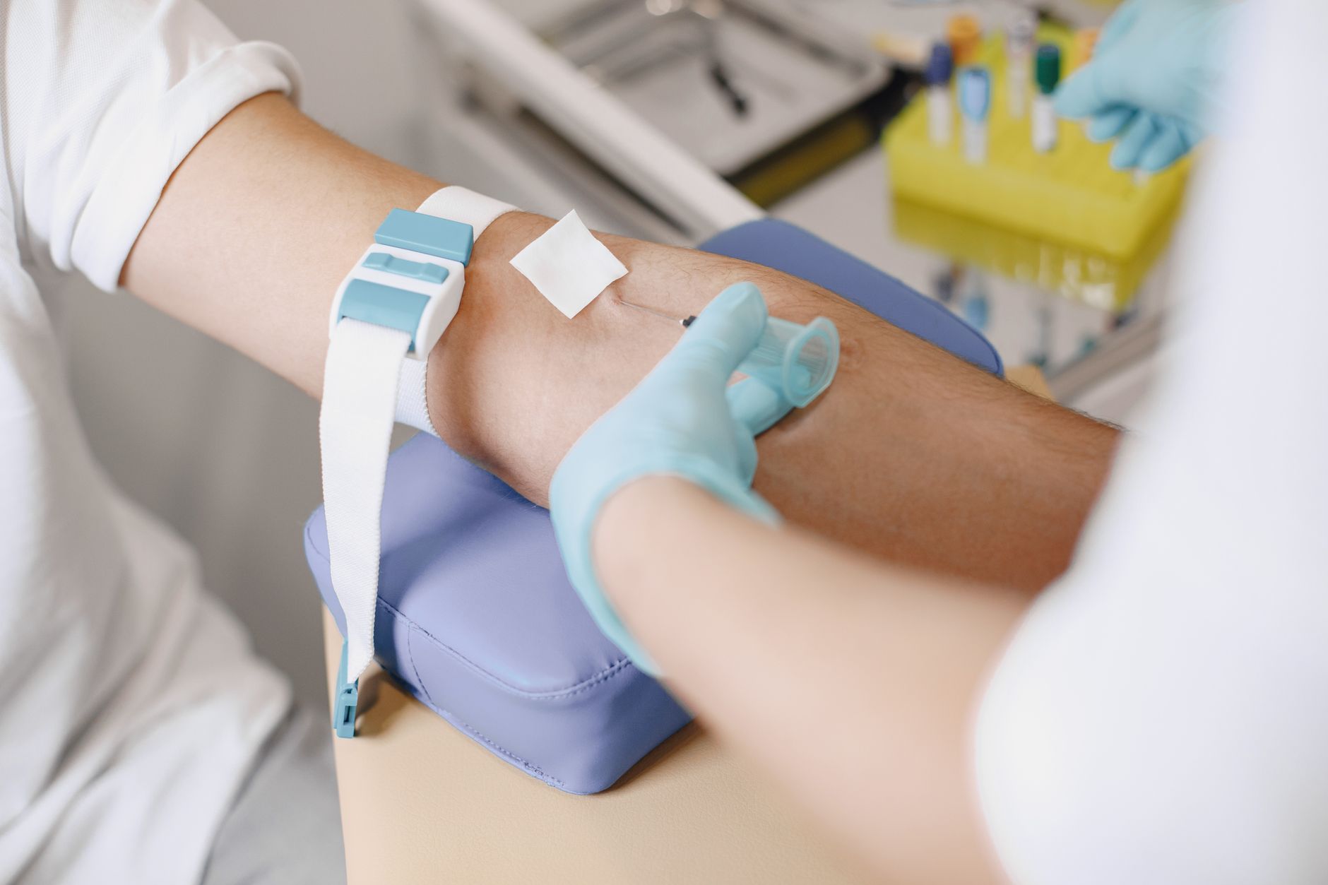 A gloved clinician draws blood from a patient's arm, with a tourniquet in place and sample tubes in a rack behind