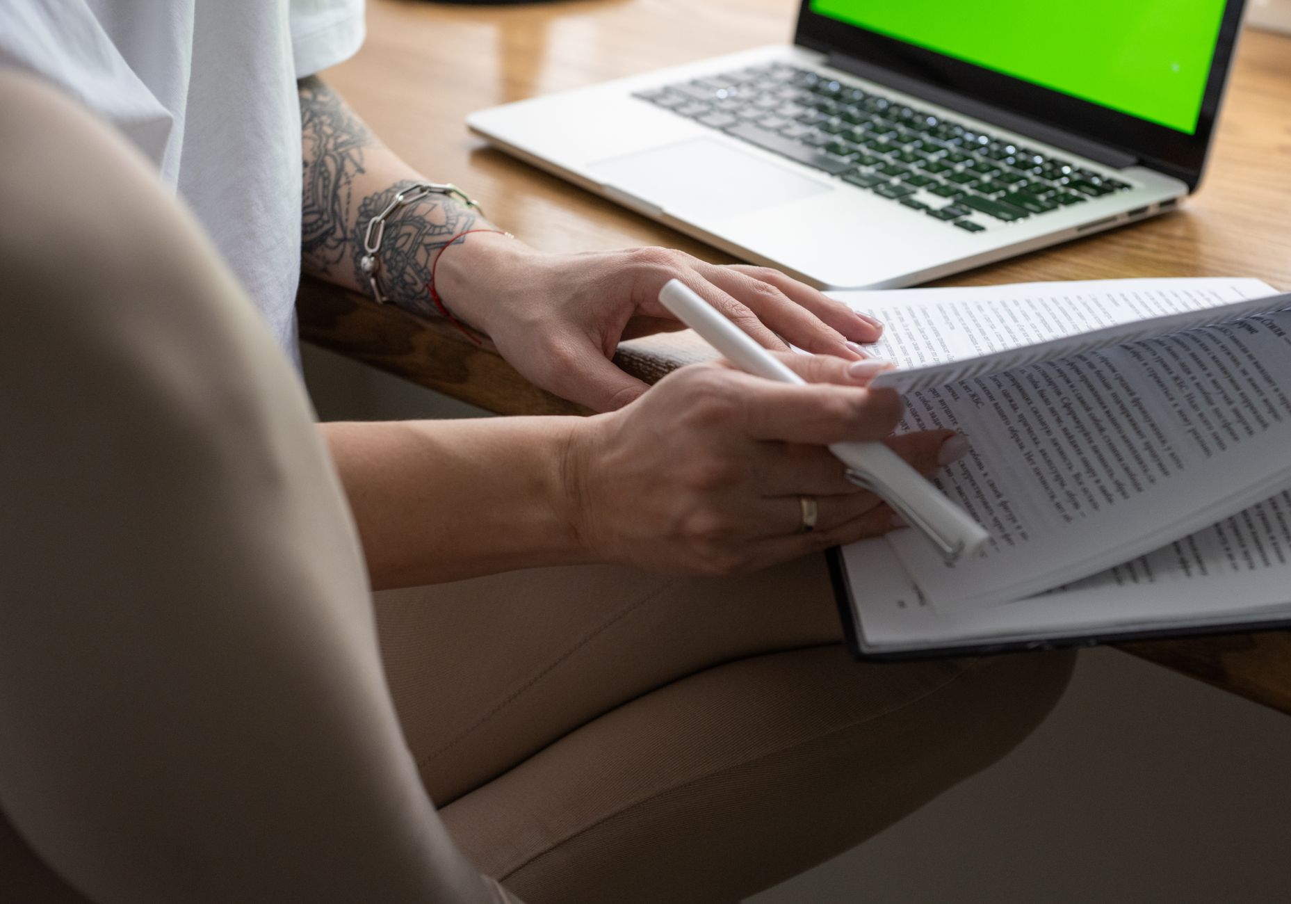 A student with a tattooed forearm reads a printed study packet with a pen in hand beside an open laptop