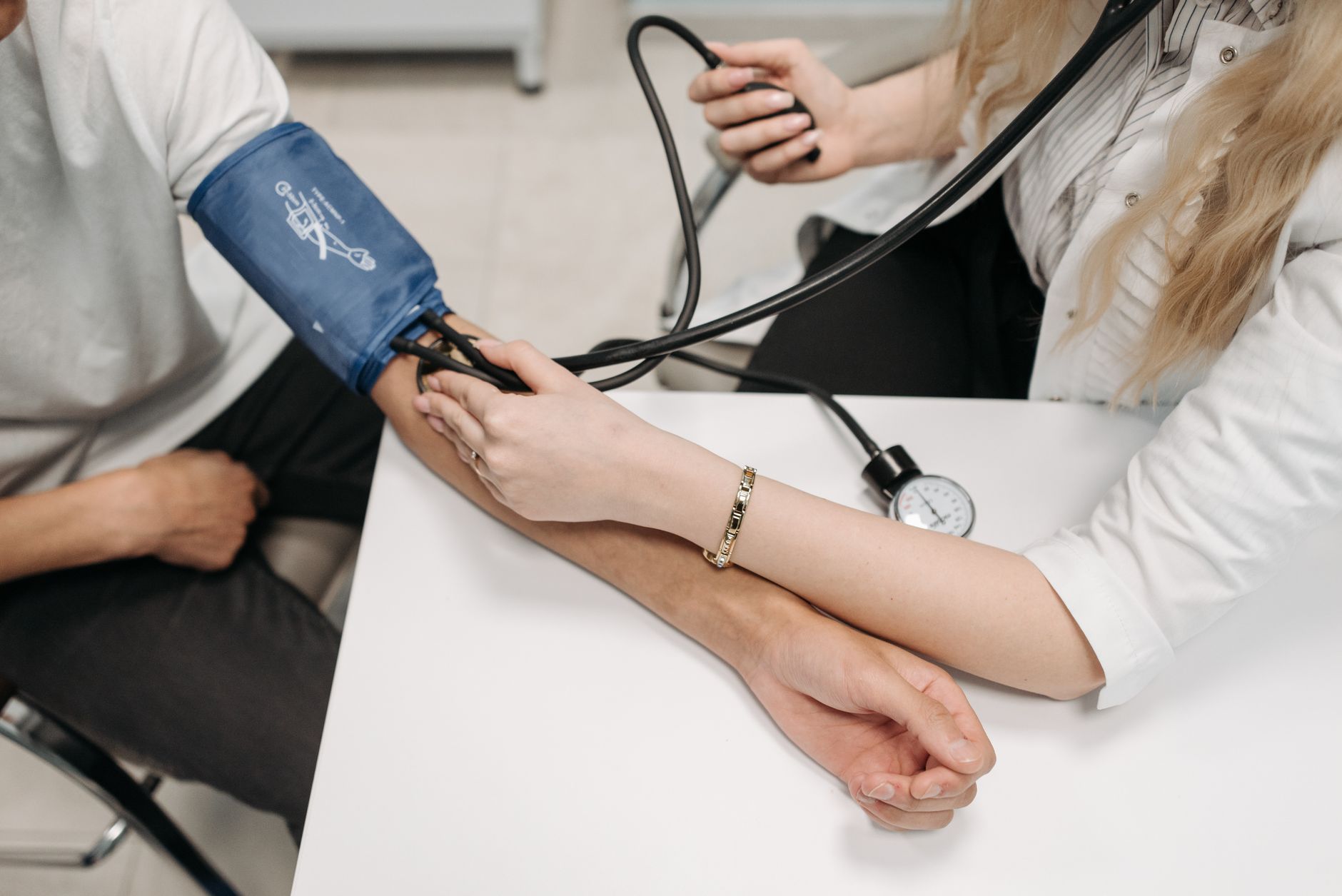 A clinician squeezes the bulb of a manual blood pressure cuff on a seated patient's arm at a white desk